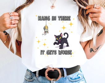 May include: White t-shirt with the text "HANG IN THERE IT GETS WORSE". The graphic features a black cat with yellow eyes and a mouse hanging from a string. Yellow star accents are scattered around the design.