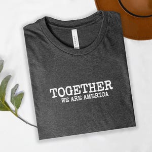 Può includere: T-shirt grigio scuro con la scritta bianca "TOGETHER WE ARE AMERICA". La maglietta è piegata, con un cappello marrone sullo sfondo. Un rametto di verde è nell'angolo in basso a sinistra.