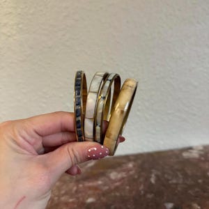 May include: Four brass bangles with inlaid designs. One bangle features dark blue rectangular inlays, two have white and iridescent inlays, and the last has a light brown, wood-like appearance. The bangles are held in a hand.