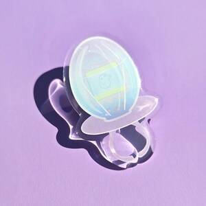 May include: A clear, iridescent pacifier-shaped pin with an egg design. The egg is light blue with green and yellow accents. The pin is on a purple background.