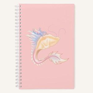 May include: A pink spiral-bound notebook with a dragon illustration. The dragon is depicted in pastel colours, with a cream head, pink body, and blue and white accents. The notebook is blank and ready for notes.