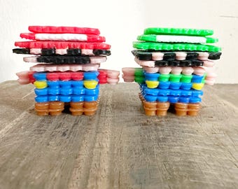 May include: Two pixel art figures, one in red and blue, the other in green and blue. The figures are made of small, colorful plastic pieces stacked to create a 3D effect. The figures are standing on a wooden surface.