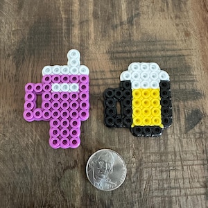 May include: Two pixel art designs: a purple drink with white accents and a black beer mug with yellow foam. A US quarter coin is next to the designs. The items are made of small, colourful beads.