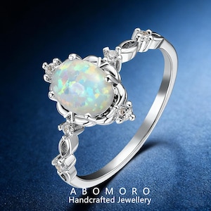 May include: Silver ring featuring an oval opal gemstone with iridescent colors. The ring is accented with small, clear gemstones and decorative silver detailing. The text "ABOMORO Handcrafted Jewellery" is at the bottom.