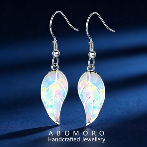 May include: A pair of silver leaf earrings with iridescent opal-like centers. The earrings have a hook closure and are displayed against a dark blue background. The text "ABOMORO Handcrafted Jewellery" is at the bottom.