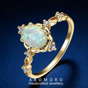 May include: A gold ring featuring an oval opal gemstone as the centerpiece, surrounded by smaller clear gemstones. The ring's band and setting are gold-colored. The text "ABOMORO Handcrafted Jewellery" is at the bottom.