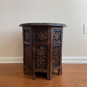May include: A dark brown, hexagonal side table with intricate carved floral designs. The table has a detailed, carved top and openwork sides, showcasing the craftsmanship. The table is made of wood and stands on short legs.
