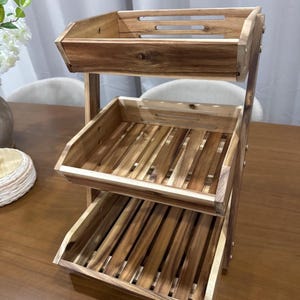 May include: A three-tiered wooden display shelf with a natural finish. The shelves are slatted, and the top shelf has a solid front. The unit is supported by vertical wooden posts and is ideal for displaying items in a kitchen or store.