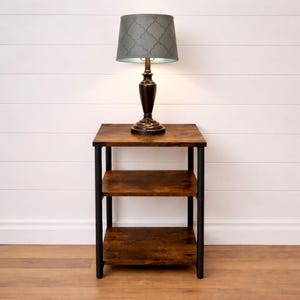 May include: A dark brown lamp with a gray patterned lampshade sits on a small, brown, three-tiered side table with black metal legs. The lamp base is a dark brown color. The table is placed against a white wall.