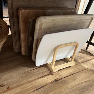 May include: A collection of wooden cutting boards of various sizes and shapes, including rectangular and square designs. A white cutting board is displayed in a bamboo holder. The boards are arranged on a wooden surface.