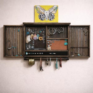 May include: A rustic wooden jewelry armoire with multiple compartments and doors, filled with necklaces, earrings, and bracelets. A yellow painting of a butterfly hangs above the armoire. The armoire has a dark brown finish.