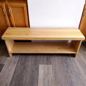 May include: A light-colored wooden bench with a lower shelf. The bench is made of natural wood and has a simple, rectangular design. The bench measures approximately 48 inches long, 14 inches wide, and 18 inches high.