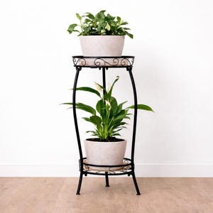 May include: A two-tiered black metal plant stand with two potted plants. The stand has a decorative scrolled design on the top and bottom tiers. The plants have green foliage and are in light-colored pots.