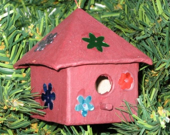 May include: A red birdhouse Christmas ornament with a triangular roof and a round entry hole. The birdhouse is decorated with colorful flower-shaped embellishments and hangs from a gold string against a backdrop of green Christmas tree branches.