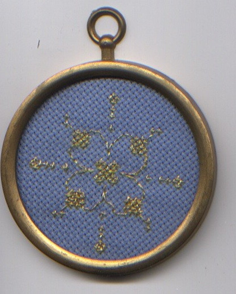 Gold stitching outlining a four-petaled dogwood flower on a medium blue background, framed in a circular old-gold toned plastic frame with a hanging loop at the top.