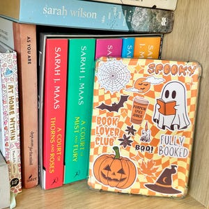 Może przedstawiać: Drewniany regał z różnymi książkami i dekoracjami. Widoczna jest obudowa telefonu z motywem Halloween z duchem i dynią, a także książki Sarah J. Maas. Na obudowie telefonu widnieje napis "Spooky".