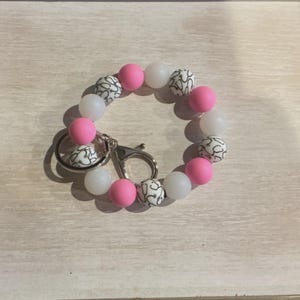 May include: A key ring bracelet featuring a circular design of pink, white, and black patterned beads. The bracelet has a silver-colored clasp and key ring attachment. The beads are smooth and evenly spaced.