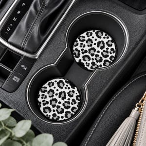 May include: Two circular car cup holder inserts featuring a black, white, and gray leopard print pattern. The coasters are positioned inside the dual cup holder console of a vehicle, adjacent to the gear shift and drive mode controls.