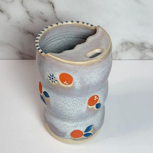 May include: A cylindrical ceramic tumbler with a speckled light gray glaze, featuring a scalloped rim and a small rectangular opening. The exterior is decorated with hand-painted orange fruit motifs and small blue floral accents around the body.