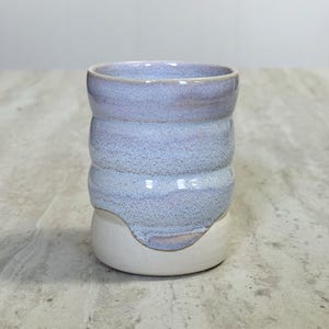 Może przedstawiać: Ręcznie robiony ceramiczny kubek z jasnoniebieskim i białym szkliwem. Kubek ma unikalny, teksturowany design z zaokrągloną podstawą i serią poziomych żeber. Wnętrze jest ciemniejszym odcieniem niebieskiego.