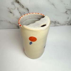 Può includere: Un bicchiere in ceramica color crema con un frutto arancione dipinto a mano e piccole foglie blu. Il bordo è decorato con un motivo a linee tratteggiate arancioni e la parte superiore include un coperchio con bordo ondulato e apertura.