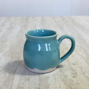 May include: A handmade ceramic mug with a rounded body and a curved handle. The mug is glazed in a glossy teal color, with a white, scalloped base. The mug is designed for hot or cold beverages.