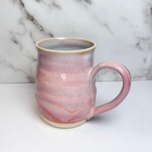 May include: A ceramic mug with a rounded, slightly textured body and a curved handle. The exterior features a glossy glaze in shades of soft pink and lavender, transitioning to a lighter, creamy off-white base. The interior is a pale grey.