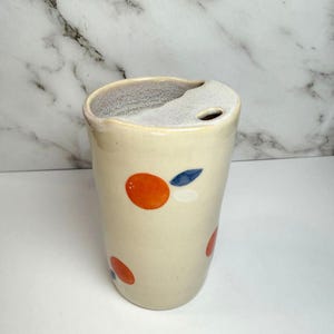 May include: A cream-colored ceramic travel tumbler with a matching lid featuring a small drinking hole. The exterior is decorated with hand-painted orange circles and small blue leaf accents. The interior rim has a textured, speckled finish.
