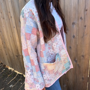 May include: A quilted jacket constructed from a patchwork of various floral and solid-colored fabric squares in shades of pink, blue, and cream. The jacket features long sleeves, an open front design, and a single patch pocket on the lower front.