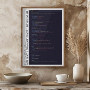 May include: A framed print featuring lines of code in a dark blue color, with the text "Code. Cry. Deploy. Repeat. HOPE IS NOT A DEPLOYMENT STRATEGY... BUT HERE WE ARE." in a vertical orientation. The frame is a light brown color.