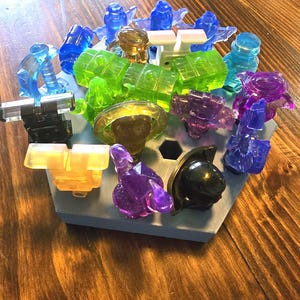 May include: A collection of colourful, translucent plastic figurines arranged on a hexagonal base. The figurines are in various shapes and sizes, including a sword, a star, a skull, and a few blocks.
