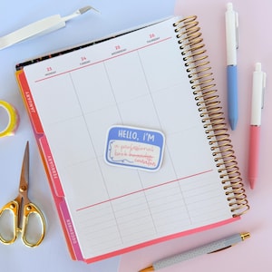 May include: A planner with a gold spiral binding, open to a blank calendar page. A sticker that says "HELLO, I'M a professional book hoarder curator" is on the page. Also, there are pens, scissors, and tape.