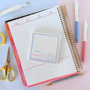 May include: A planner with a gold spiral binding, open to a week view, with a square notepad featuring a stack of books. Two pens, one blue and one pink, and a glitter pen are next to the planner. Gold scissors and washi tape are also visible.