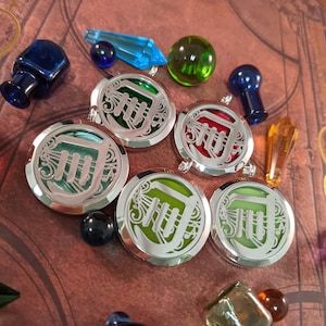 May include: A collection of silver-colored pendants with a stylized letter design, each with a different colored background. The image also includes various glass beads, crystals, and small bottles in shades of blue, green, and orange, set against a patterned background.