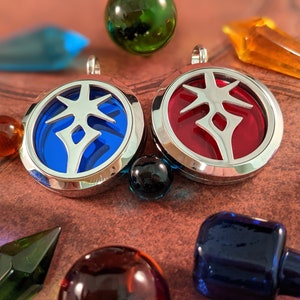 May include: Two silver-toned pendants with a circular design, one with a blue center and the other with a red center. Each pendant features a stylized emblem. The pendants are surrounded by colorful glass beads and crystals on a textured brown surface.