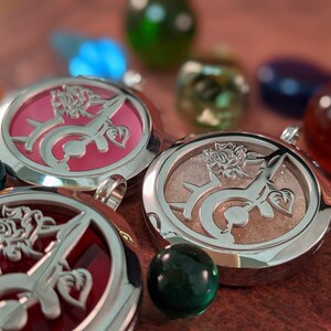 May include: Close-up of three round silver pendants with rose and vine designs. The pendants have pink, red, and tan backgrounds. Colorful glass marbles and gems surround the pendants, creating a decorative display.
