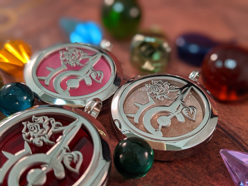 May include: Close-up of three round silver pendants with rose and vine designs. The pendants have pink, red, and tan backgrounds. Colorful glass marbles and gems surround the pendants, creating a decorative display.