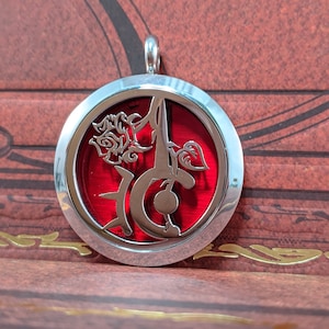 May include: A round, silver-colored pendant with a red center. The pendant features a stylized floral design and a decorative letter. The pendant is suspended from a silver loop.