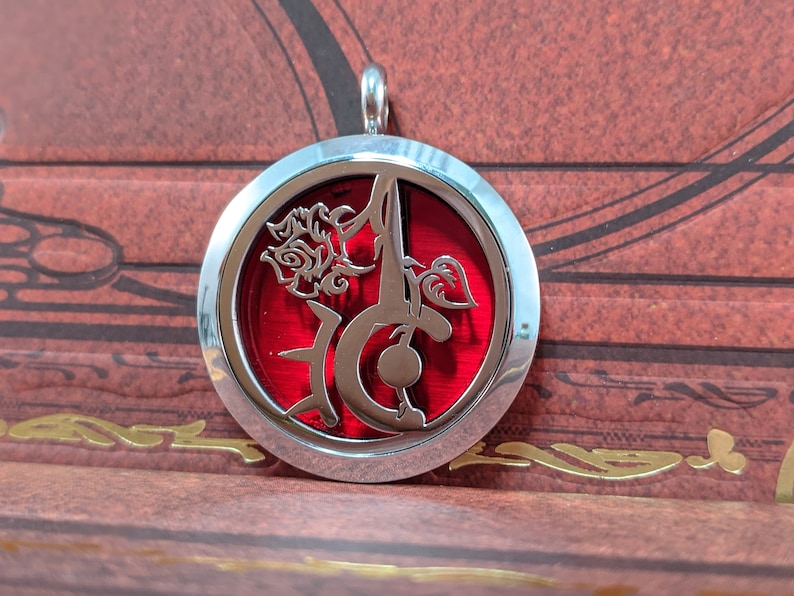 May include: A round, silver-colored pendant with a red center. The pendant features a stylized floral design and a decorative letter. The pendant is suspended from a silver loop.