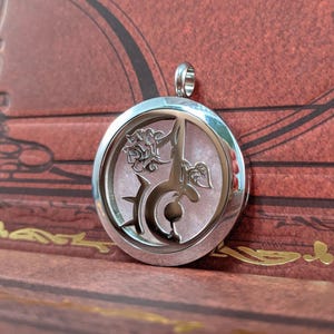 May include: A silver-colored pendant with a circular design. The pendant features a stylized rose, a sword, and other decorative elements. The background within the pendant has a pink, glittery appearance. The pendant has a loop at the top for a chain.