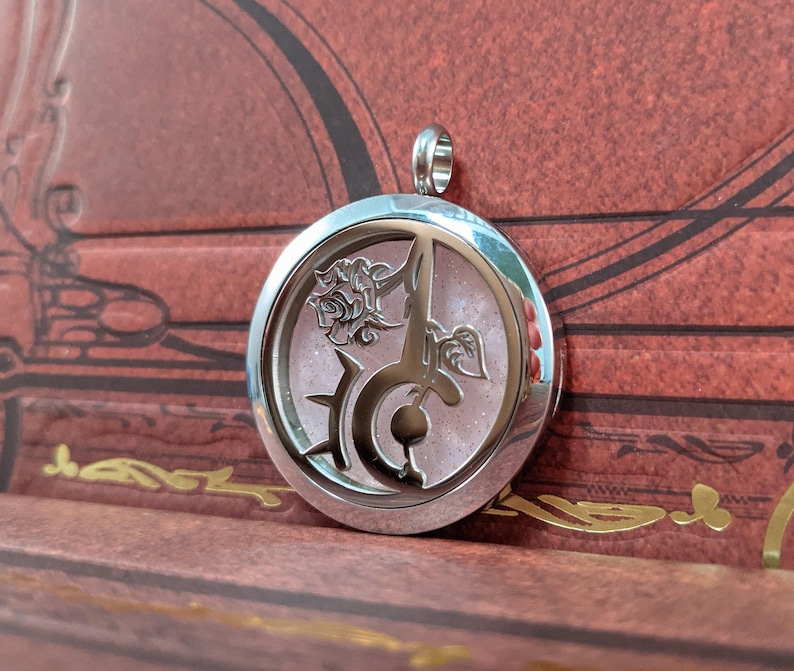May include: A silver-colored pendant with a circular design. The pendant features a stylized rose, a sword, and other decorative elements. The background within the pendant has a pink, glittery appearance. The pendant has a loop at the top for a chain.