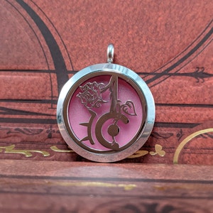 May include: A silver-colored pendant with a circular design. The pendant features a pink interior and a cut-out floral and letter design. The pendant hangs from a small silver loop.