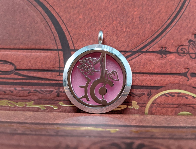 May include: A silver-colored pendant with a circular design. The pendant features a pink interior and a cut-out floral and letter design. The pendant hangs from a small silver loop.