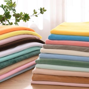 May include: Stacks of colorful fabric in various shades, including yellow, orange, pink, brown, white, teal, and blue. The fabric appears soft and plush, likely velvet or a similar material. The stacks are neatly arranged, suggesting a textile shop or craft supply store.