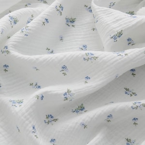 Op de afbeelding: Witte katoenen stof met een klein blauw bloemenpatroon. De stof heeft een textuur, vergelijkbaar met een wafel.