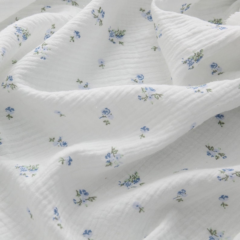 Op de afbeelding: Witte katoenen stof met een klein blauw bloemenpatroon. De stof heeft een textuur, vergelijkbaar met een wafel.