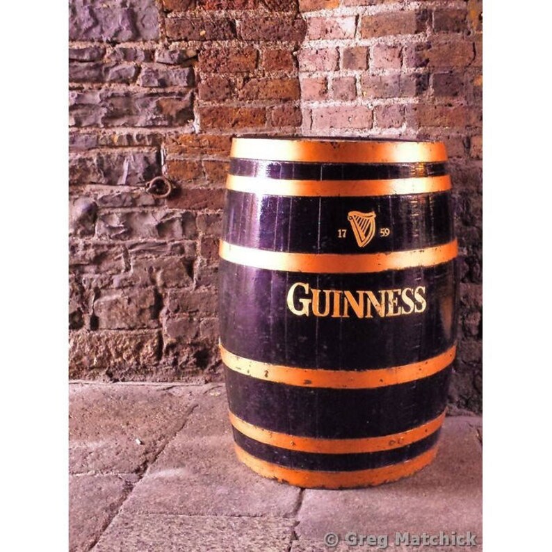 K&ouml;nnte beinhalten: Ein schwarzes Guinness-Bierfass mit goldenen Verzierungen und dem Guinness-Logo. Das Fass steht auf einem Ziegelboden.