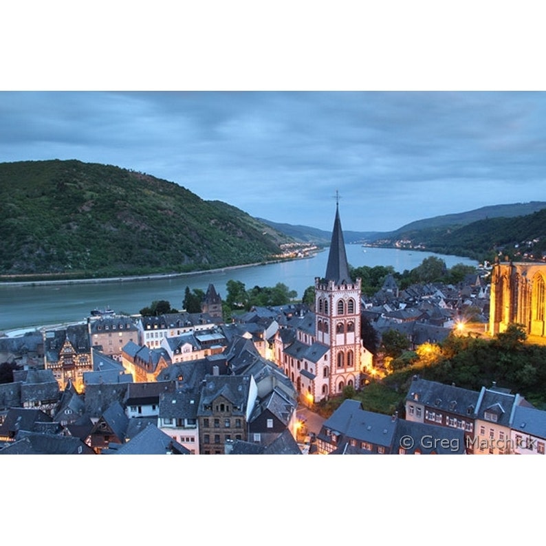 May include: A view of a small town in Germany at dusk. The town is situated on a bend in a river, with a large church in the center. The church has a tall spire and is lit up by streetlights. The town is surrounded by hills, and the sky is a soft blue.