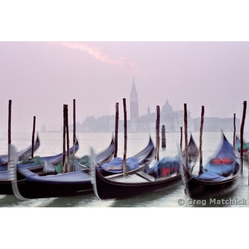May include: A misty morning scene in Venice, Italy, with several gondolas moored at a dock. The gondolas are dark blue and black, and the water is a light blue. The city skyline is visible in the background, with a tall bell tower.