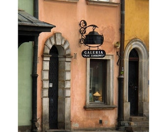 May include: A storefront with a black sign that reads "Galeria "Plac Zamkowy"". The storefront is painted a light peach color and has a black metal awning. The storefront is located on a cobblestone street.